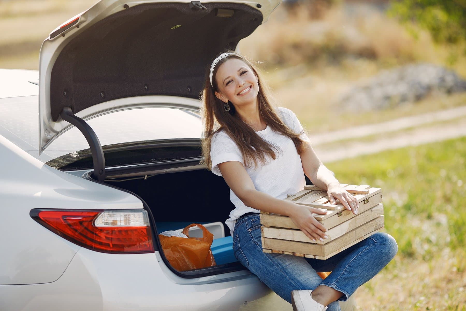 A smiling woman sitting in the open trunk of a clean white car, a wooden crate on her lap