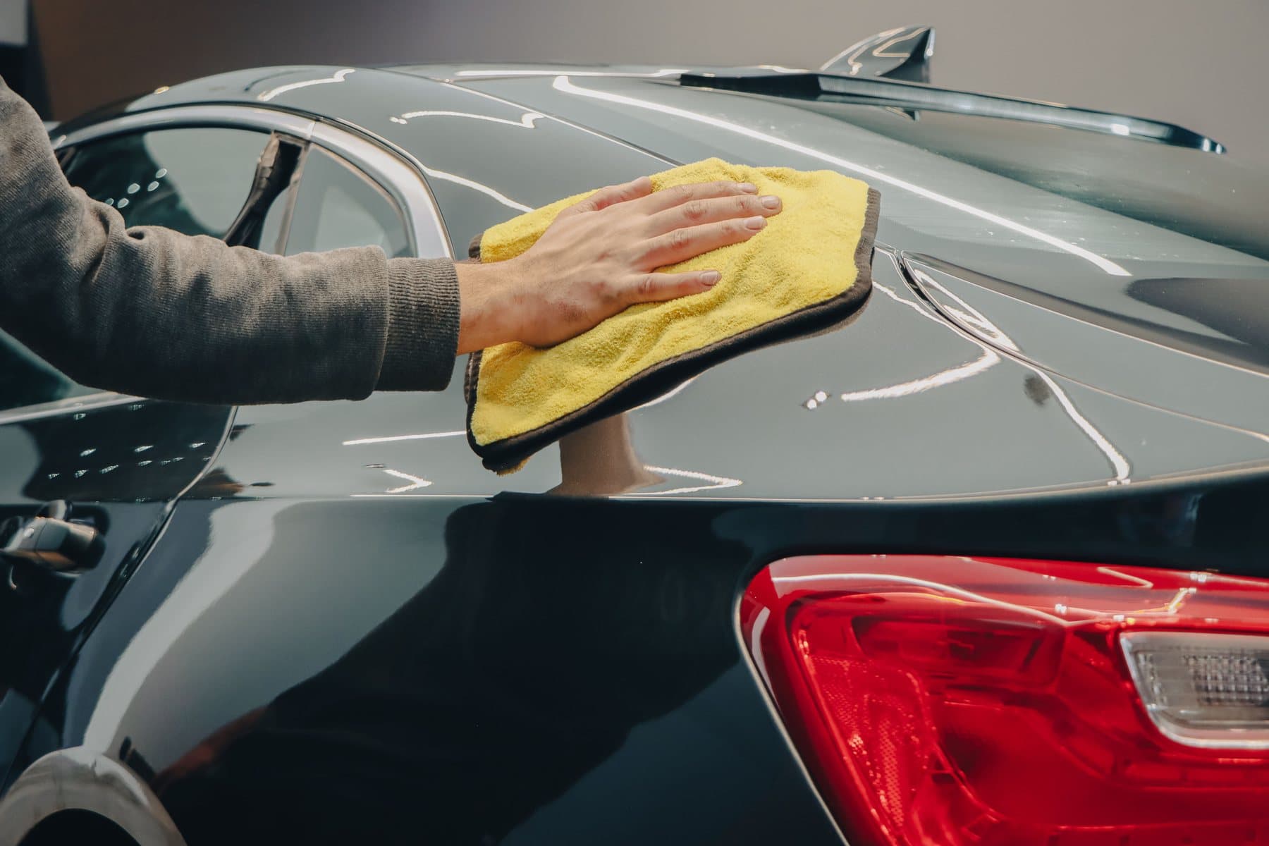 A hand wiping a glossy dark grey rear quarter panel with a yellow microfibre towel, tail lamp in the foreground