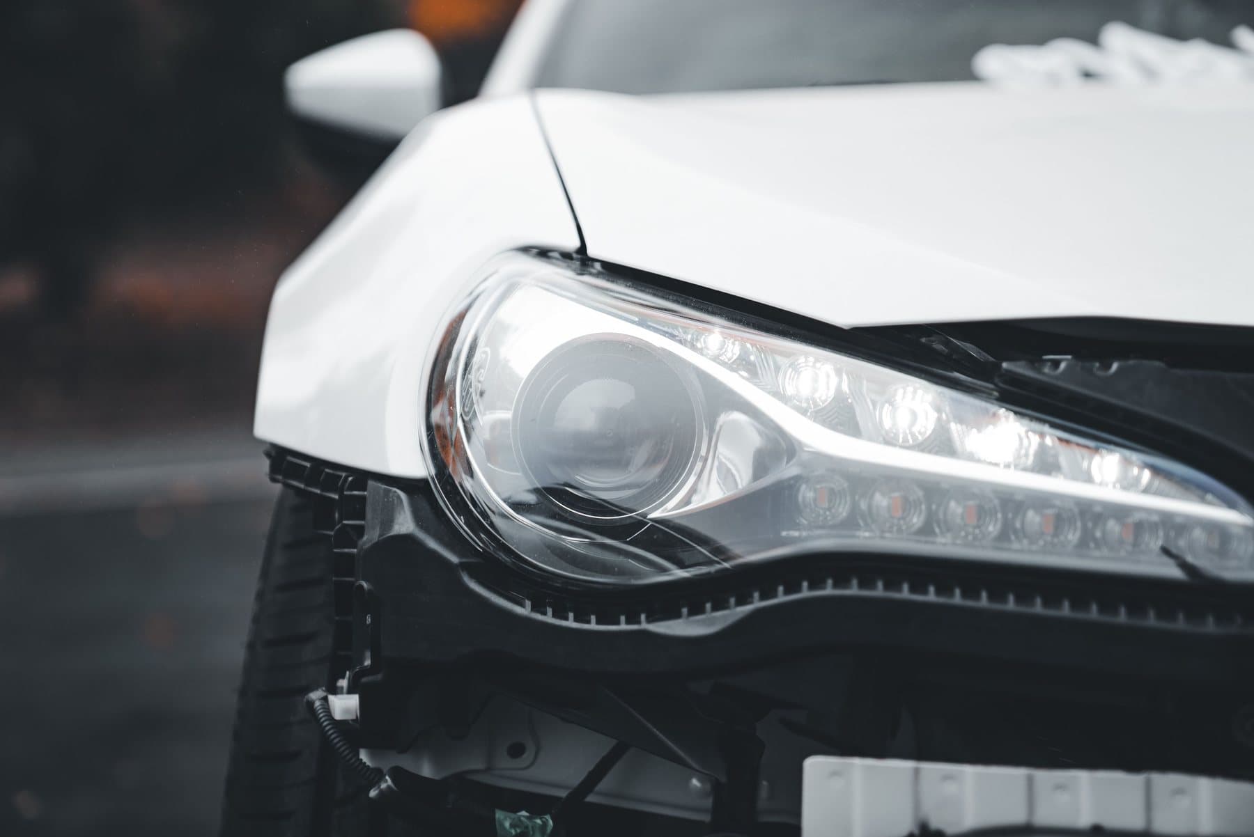 The front corner of a white car with the bumper cover damaged and pulled away below the headlight