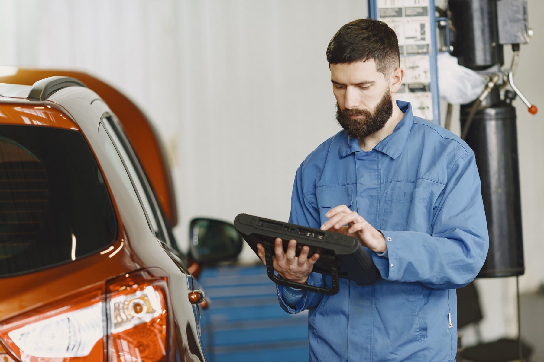 A technician in blue workwear reading a rugged tablet beside an orange car in a workshop