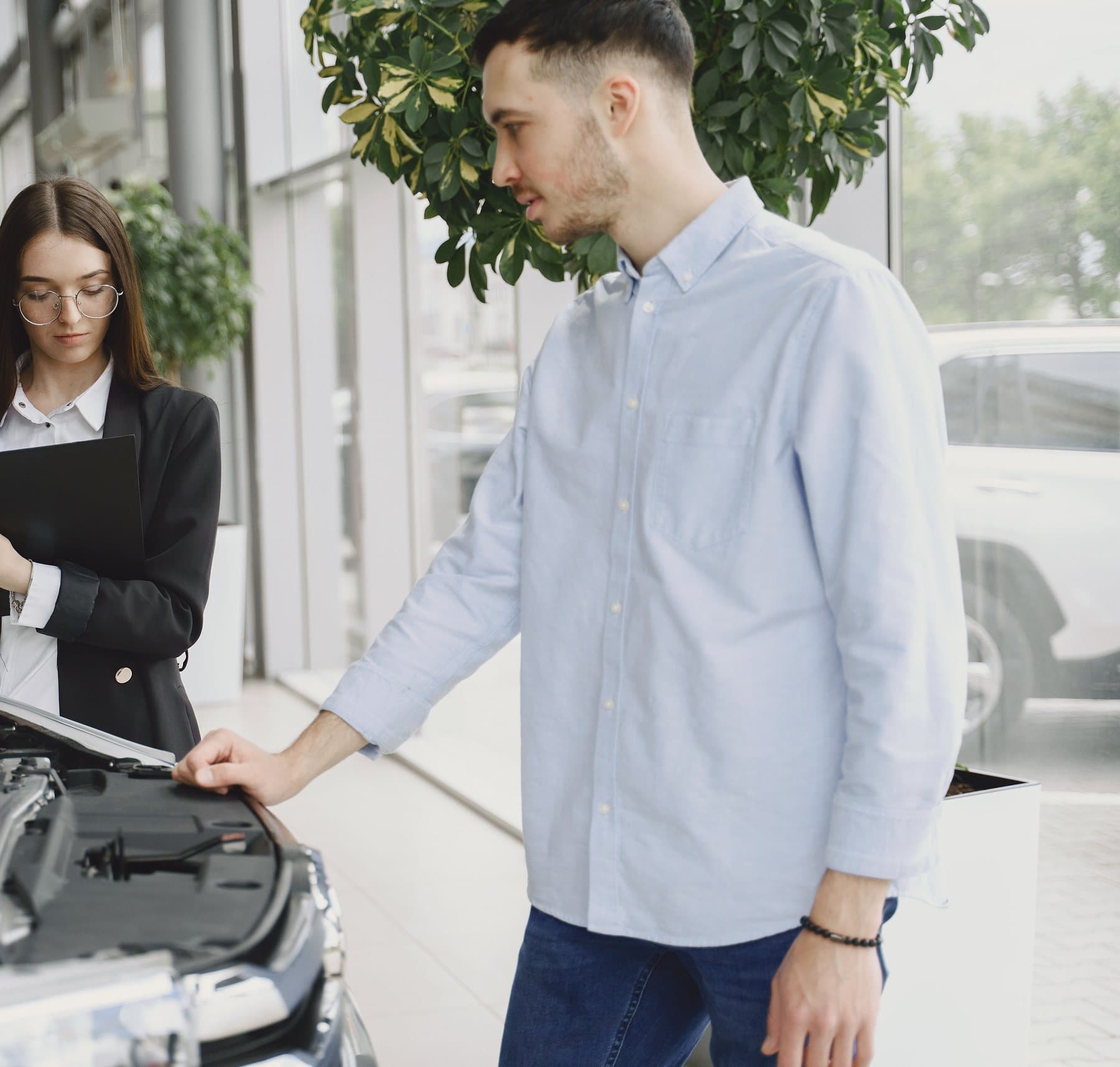 A man in a light-blue shirt, a hand on an open hood, explaining something to a woman holding a clipboard