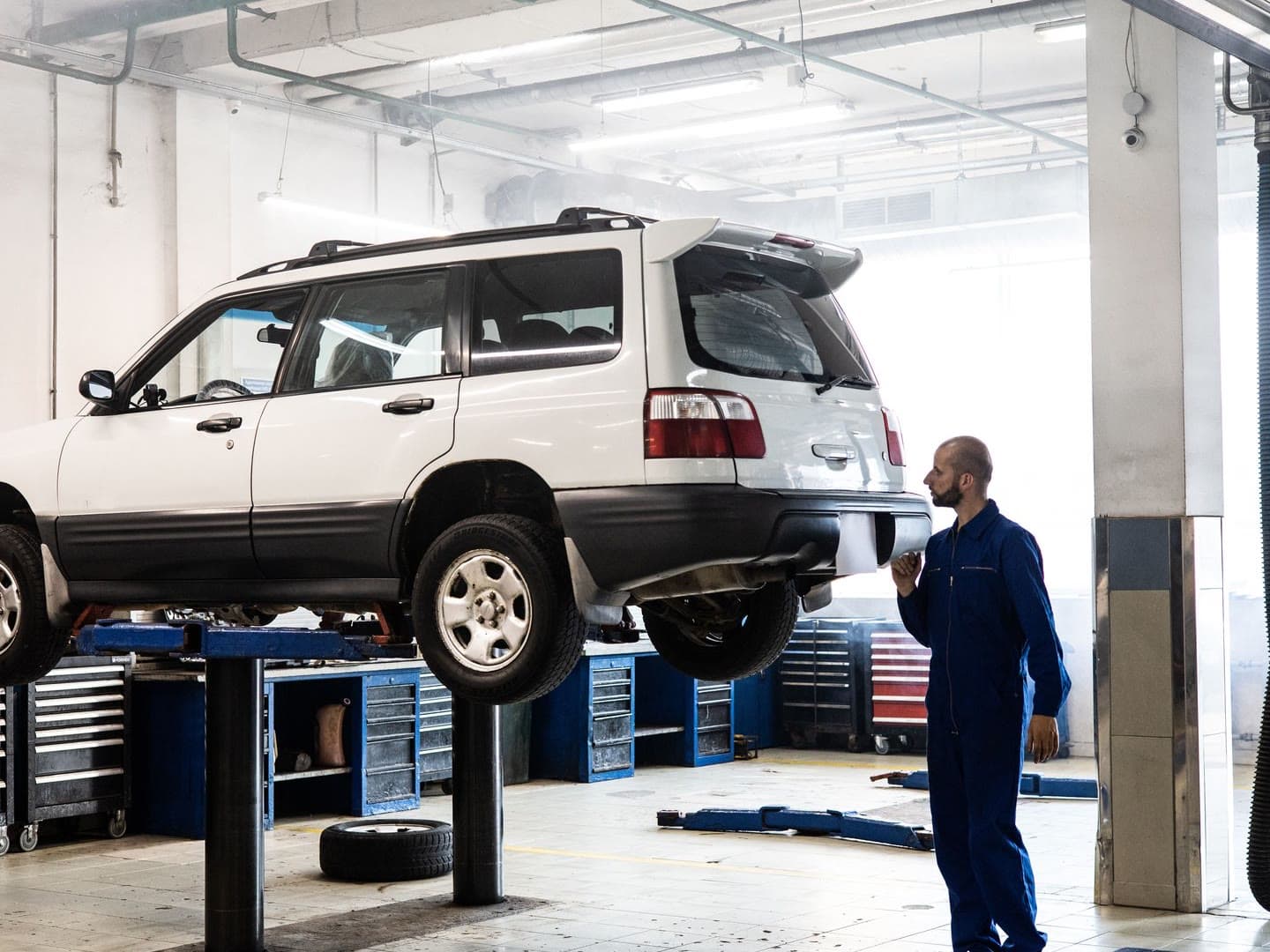A technician standing beside an SUV raised on a two-post lift in a daylit repair bay