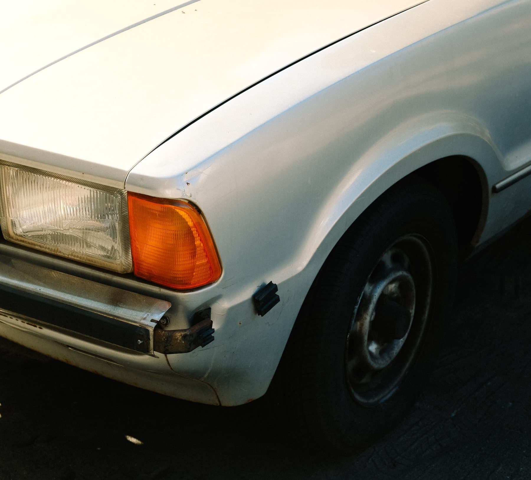 The scuffed, knocked front bumper corner of an older white car, parked in the sun