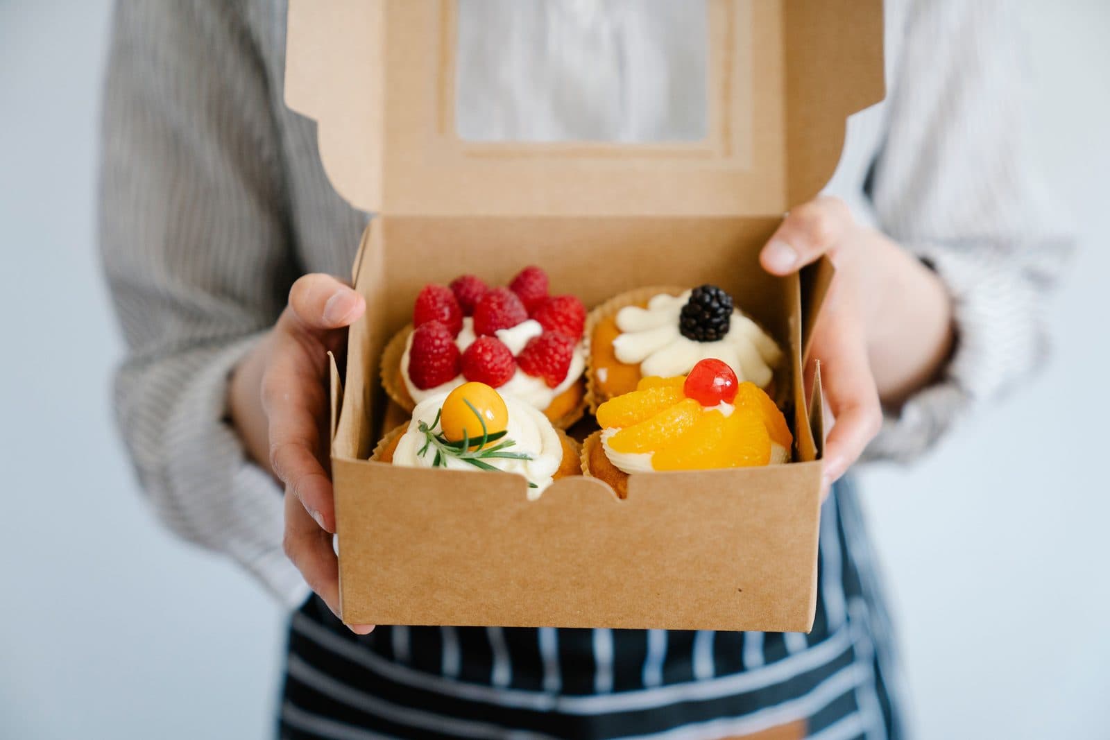 A baker in a striped apron holding an open kraft box of fruit tartlets. A photo from Sugar & Crumb Bakery, the sample bakery website.