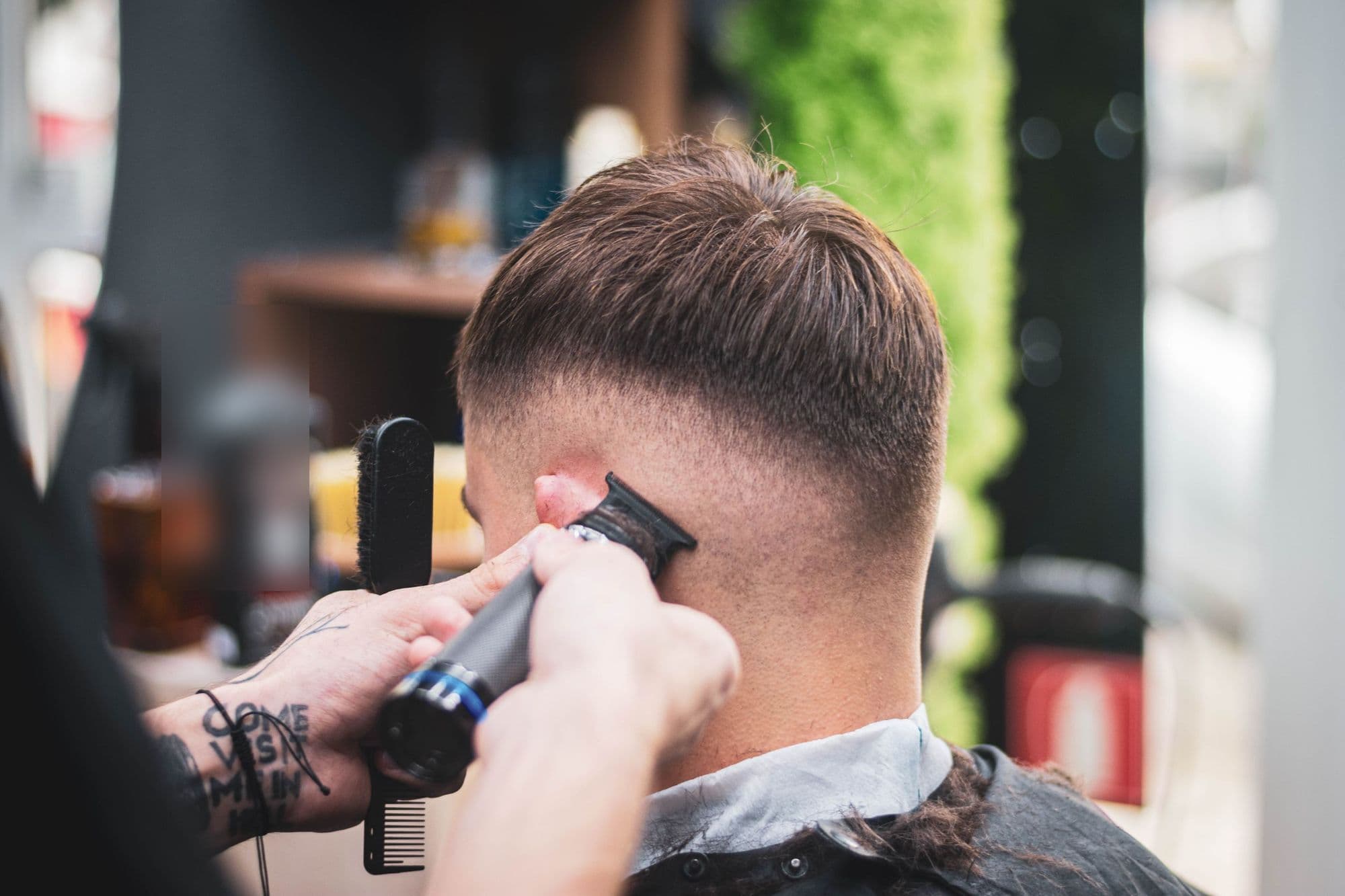 A barber tapering the back of a client’s neck with a trimmer, comb and brush in hand