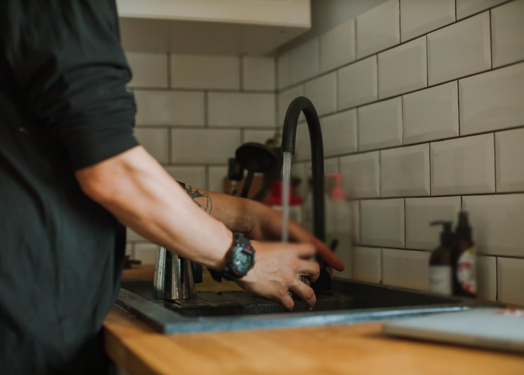 A man in a dark work shirt at a home kitchen sink, hands at the tap with the water running