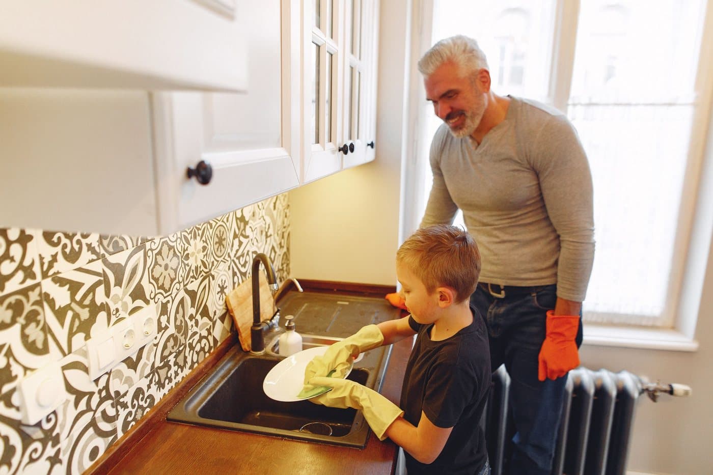 A dad and his young son washing dishes at the kitchen sink, both smiling. A photo from Benchmark Drain & Sewer, the sample drain cleaning website.