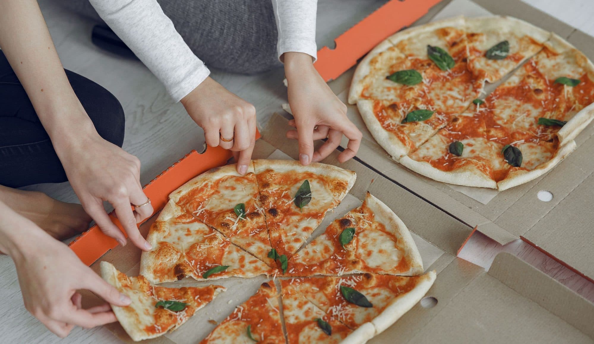 Several hands reaching for slices of two cheese and basil pizzas in open boxes, seen from above. A photo from Tomato Hill Pizzeria, the sample restaurant website.