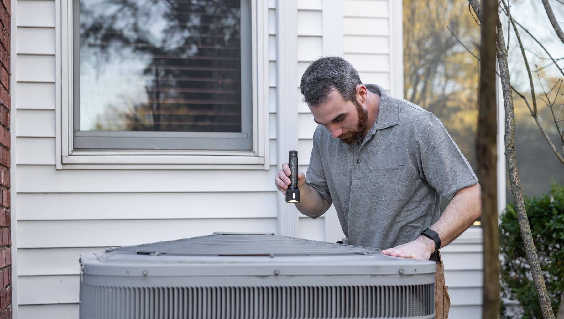 A technician with a flashlight inspecting the top of an outdoor AC condenser beside a white-sided house