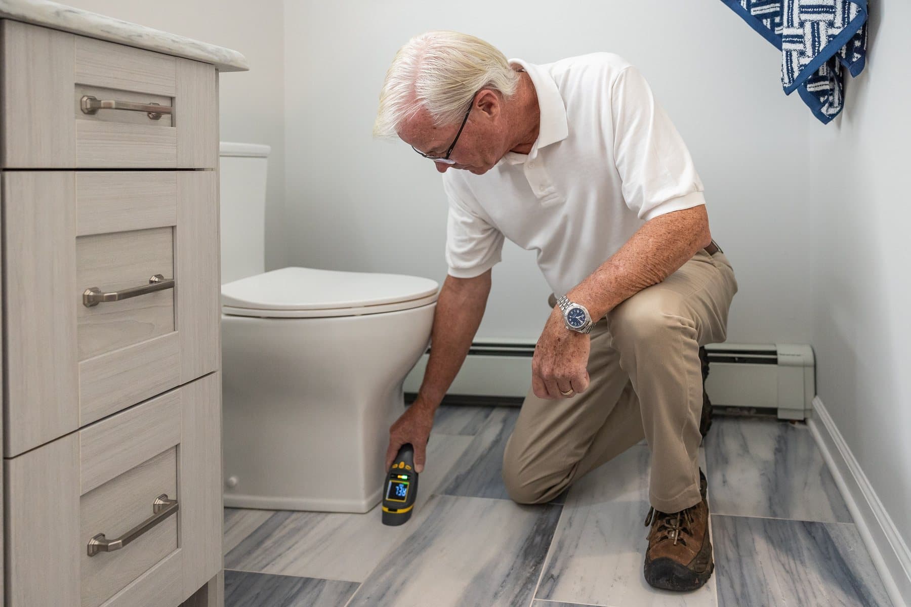 A plumber kneeling beside a toilet in a bright bathroom, holding a moisture meter to the floor
