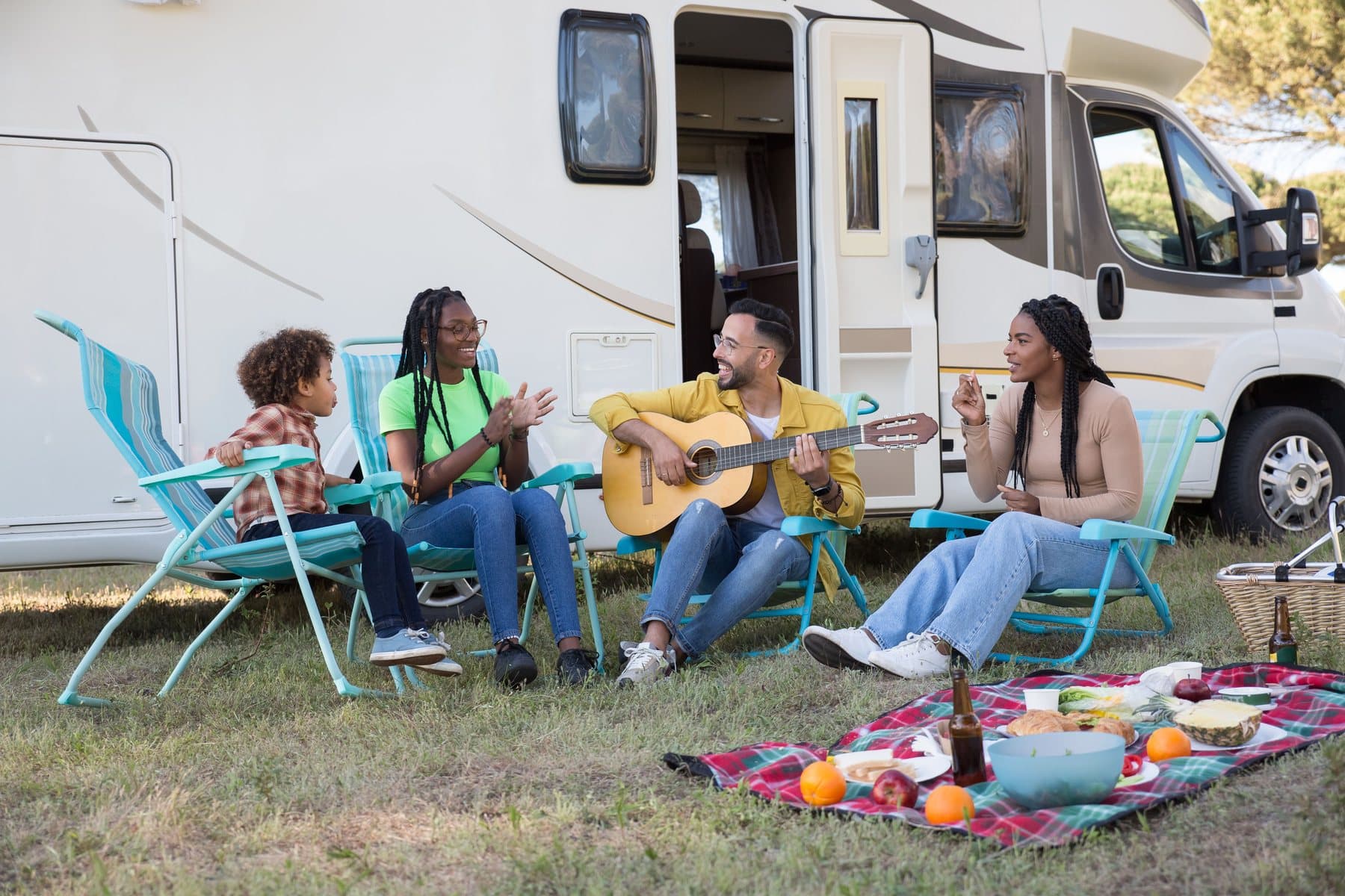 A family on camp chairs beside their motorhome, one playing guitar, a picnic spread on a blanket