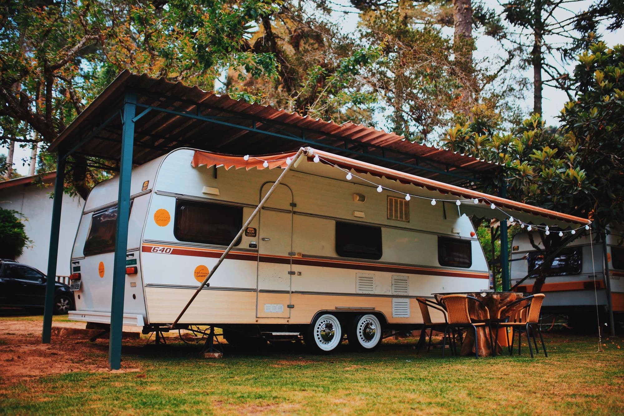 A tandem-axle travel trailer with its awning out under a carport, string lights along the edge
