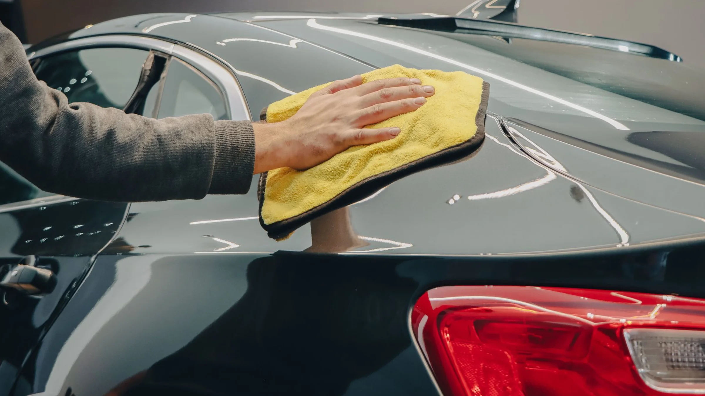 A hand wipes a glossy dark grey rear quarter panel with a yellow microfibre towel, the tail lamp in the foreground.