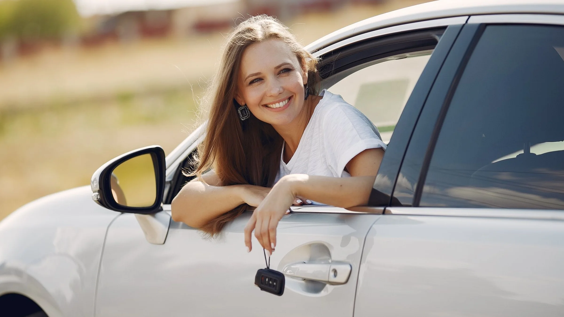 A smiling driver leans out of the window of a clean white car in the sun, a key dangling from her hand, a field behind.