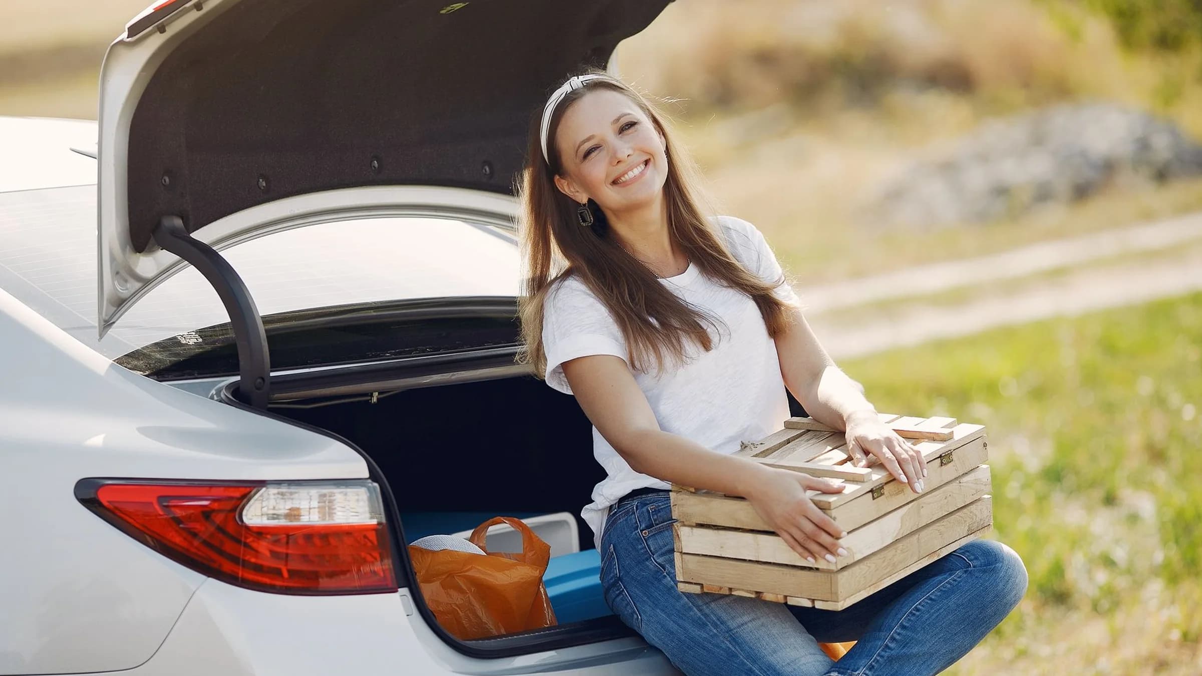 A smiling woman sits on the open trunk lip of a clean white car outdoors, a wooden crate on her lap, grass behind her.