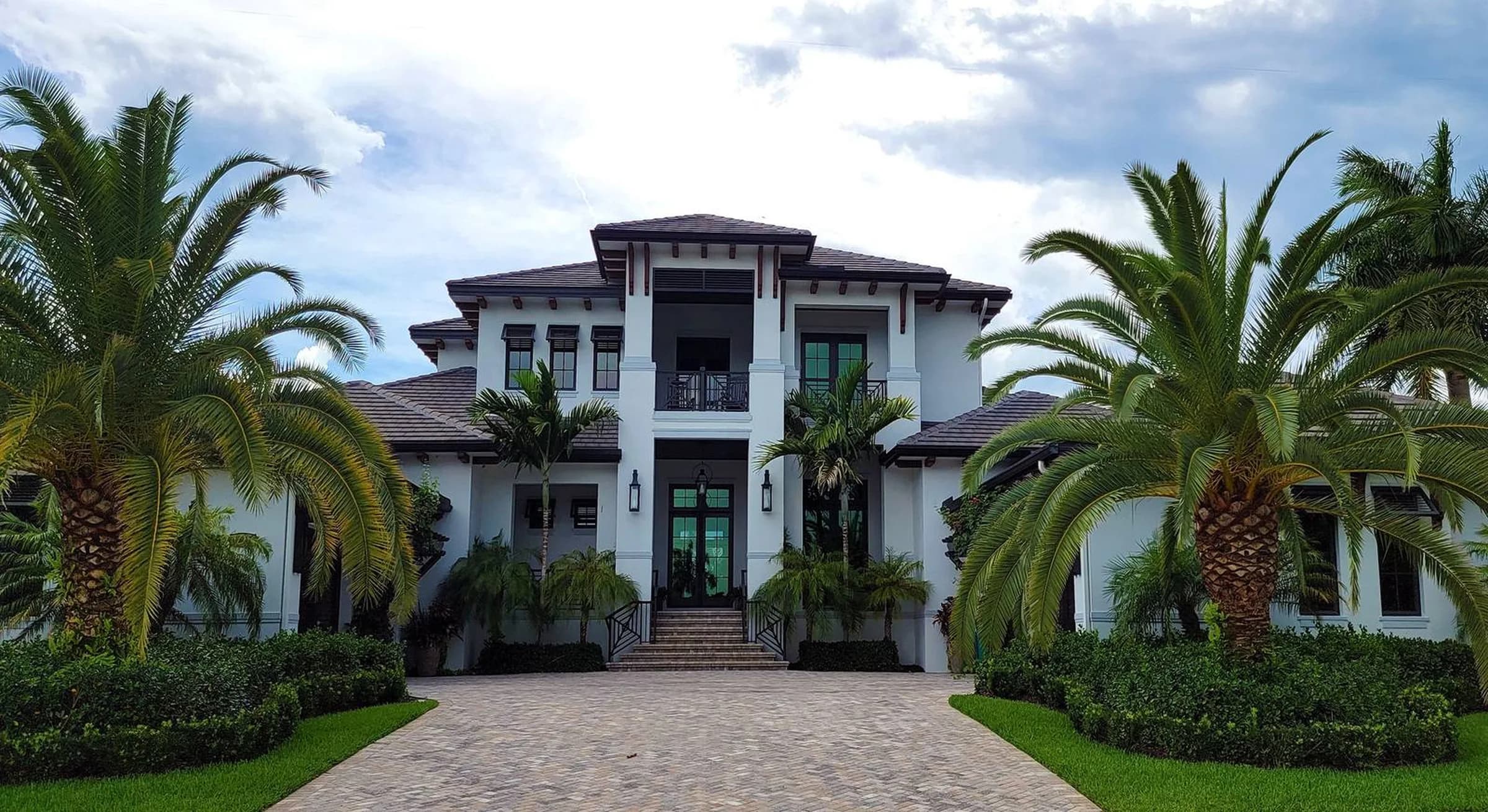 A white two-story Florida home behind a wide paver driveway, between two large palms under a partly cloudy sky.