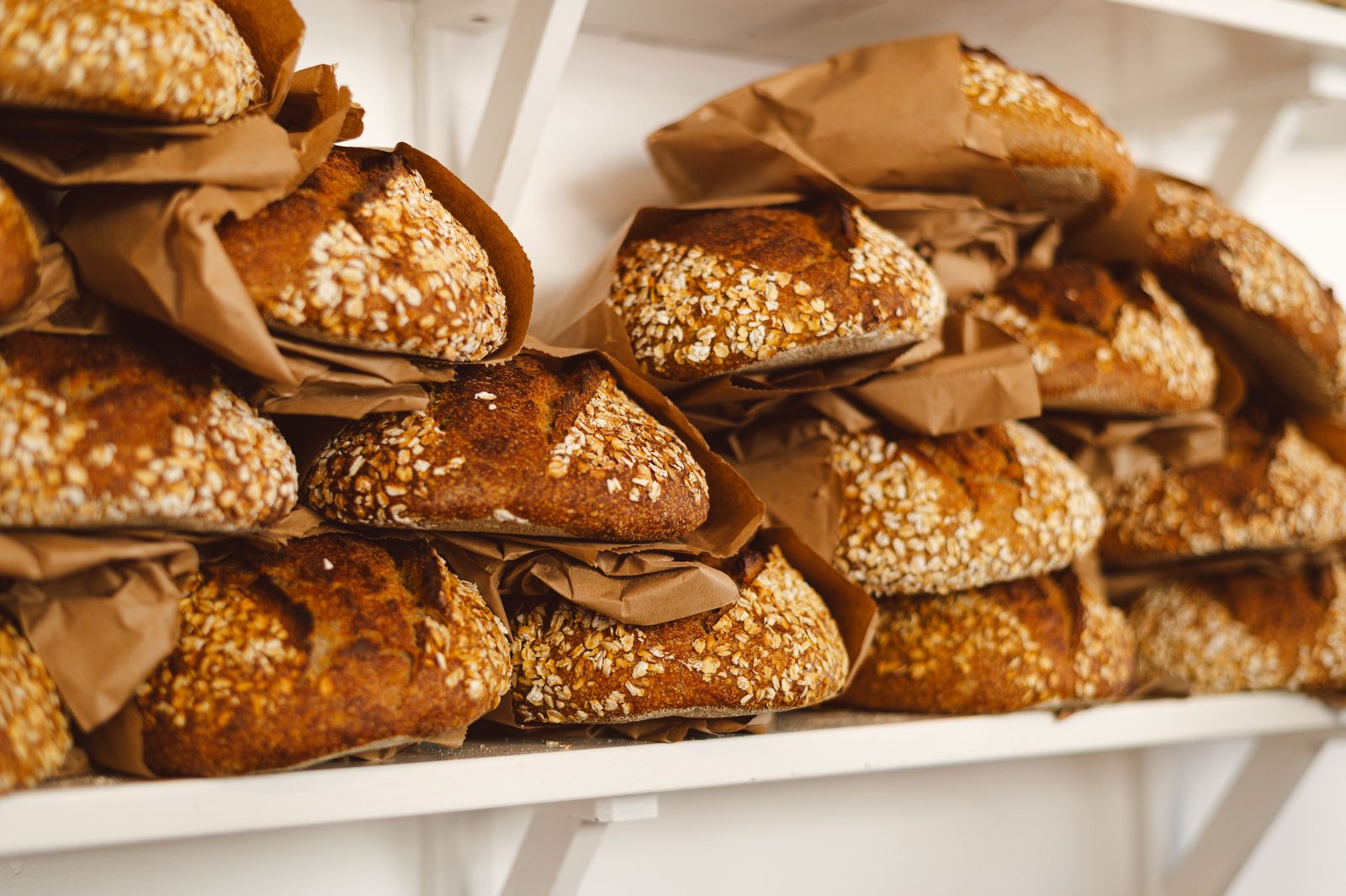 Stacked oat-crusted sourdough loaves wrapped in kraft paper on a white shelf