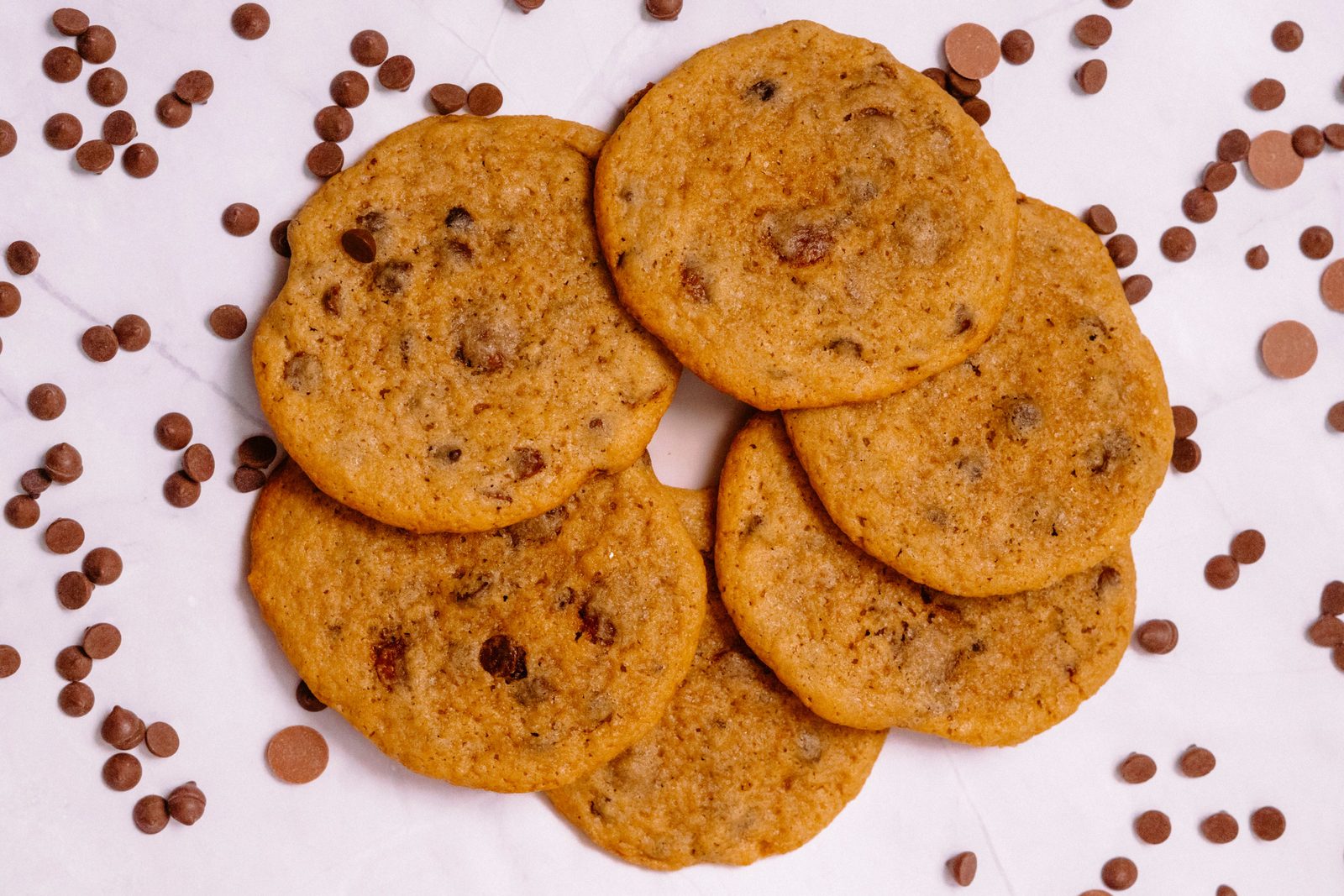 Six chocolate chip cookies on a light marble surface with scattered chocolate chunks