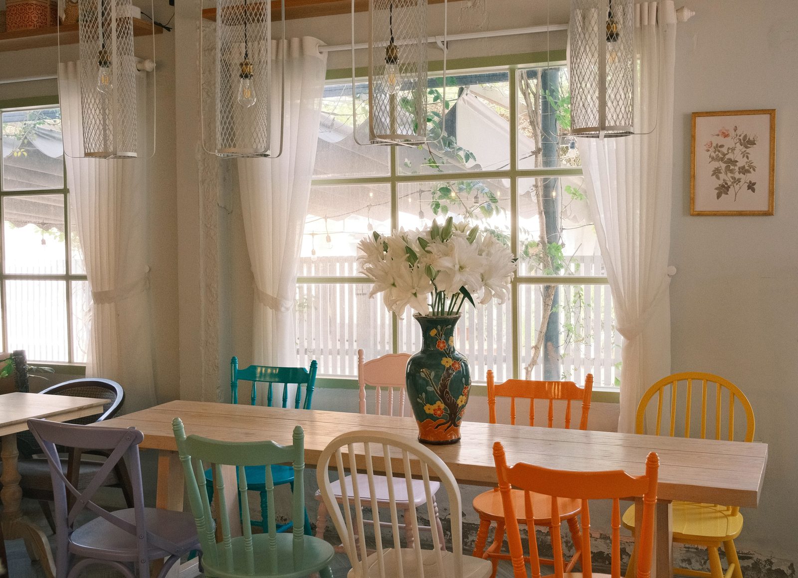 A bright cafe room with a long wooden table and mint, lilac, pink and yellow chairs