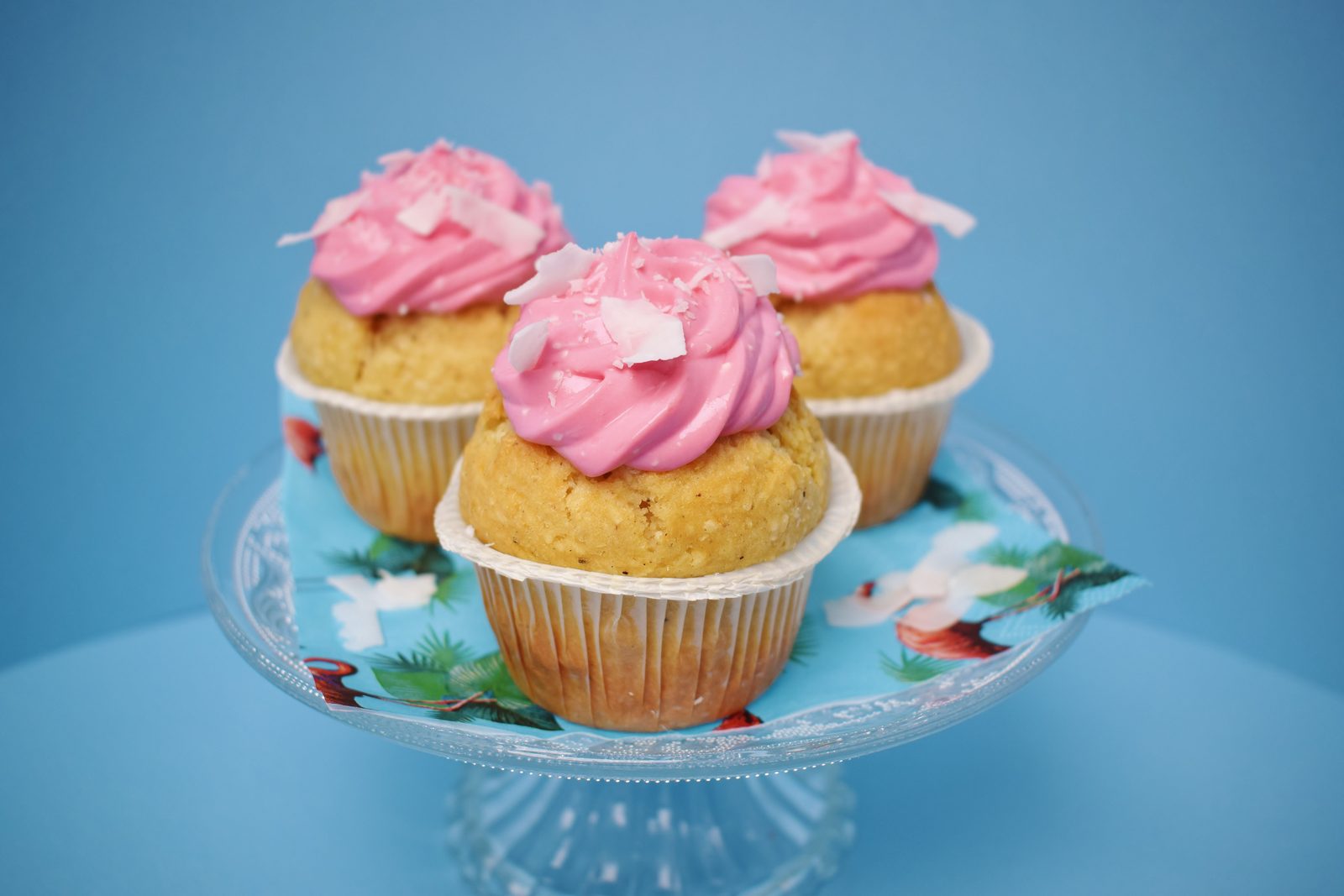Three cupcakes with swirled pink frosting and coconut flakes on a glass cake stand