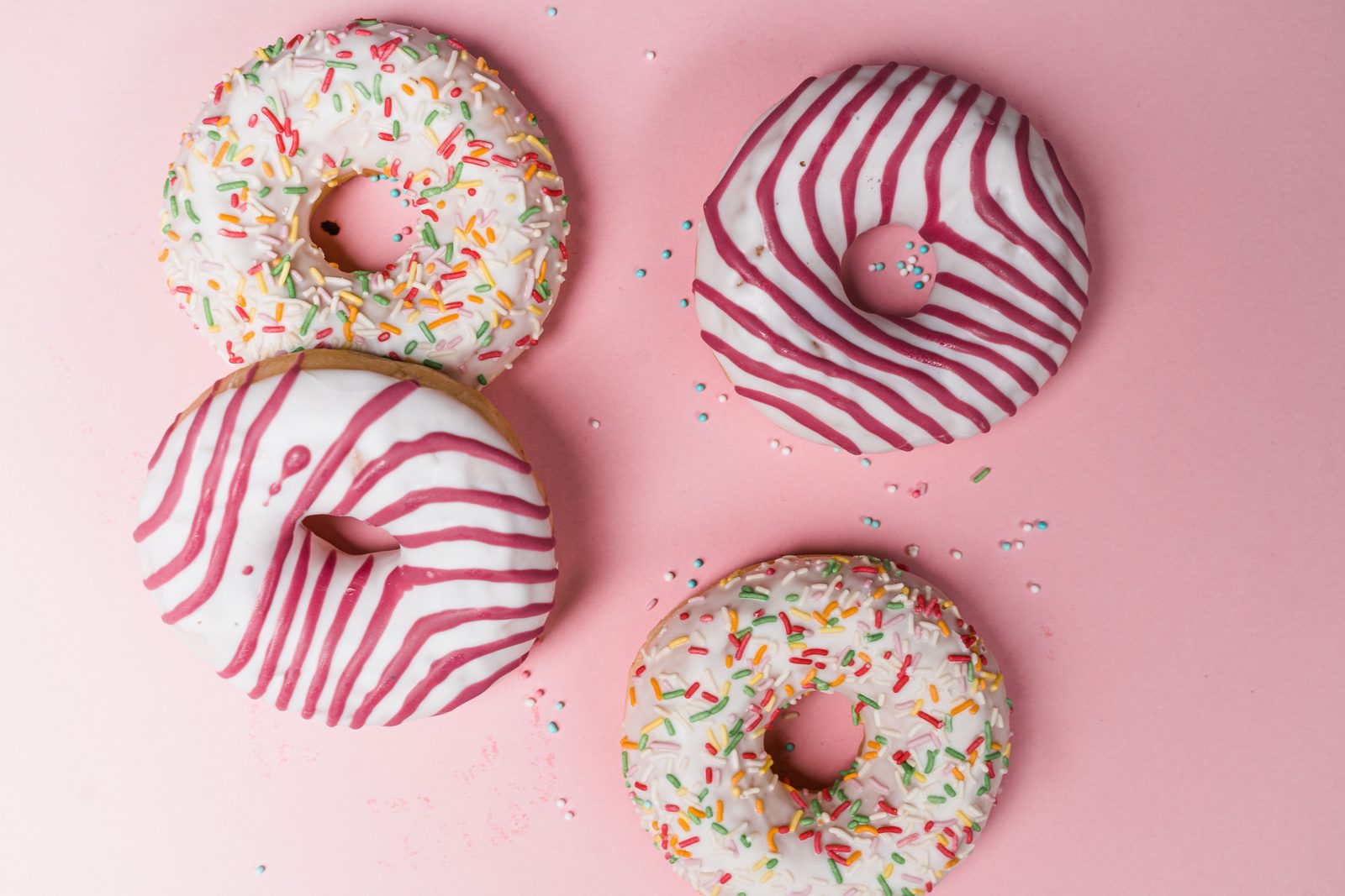 Four glazed donuts, two with rainbow sprinkles and two with raspberry stripes, on a pink table