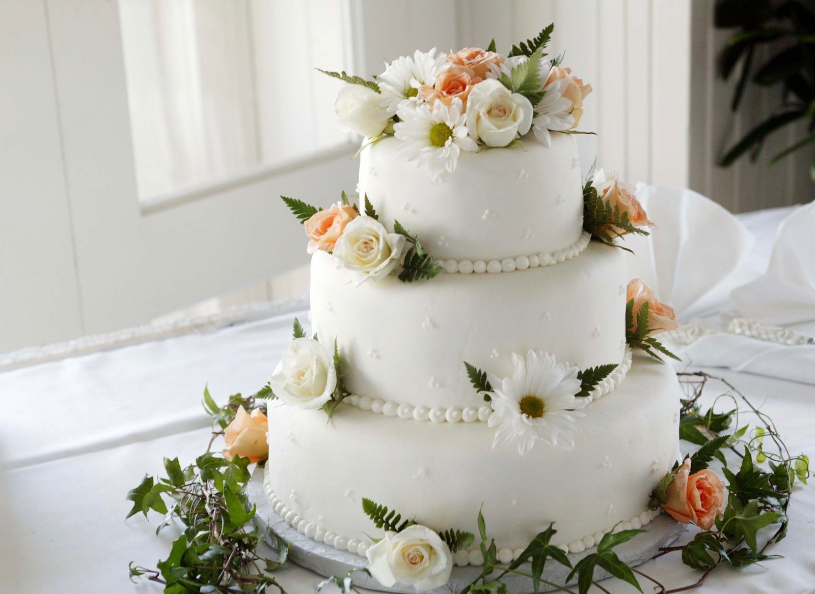 A three-tier white wedding cake with pearl piping, peach roses and daisies