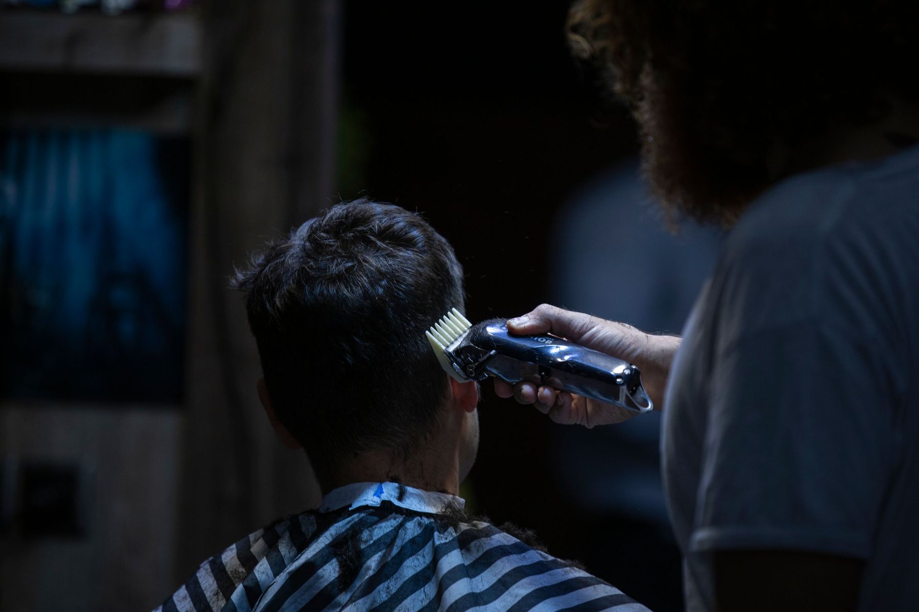 A young client in a striped cape, seen from behind while the barber runs clippers up the back