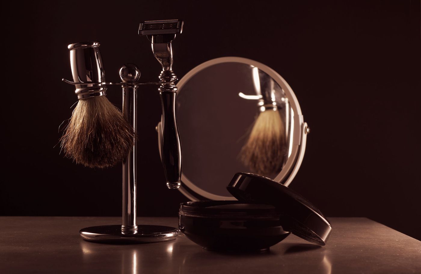A shaving brush and razor on a stand beside a mirror and a tin of soap