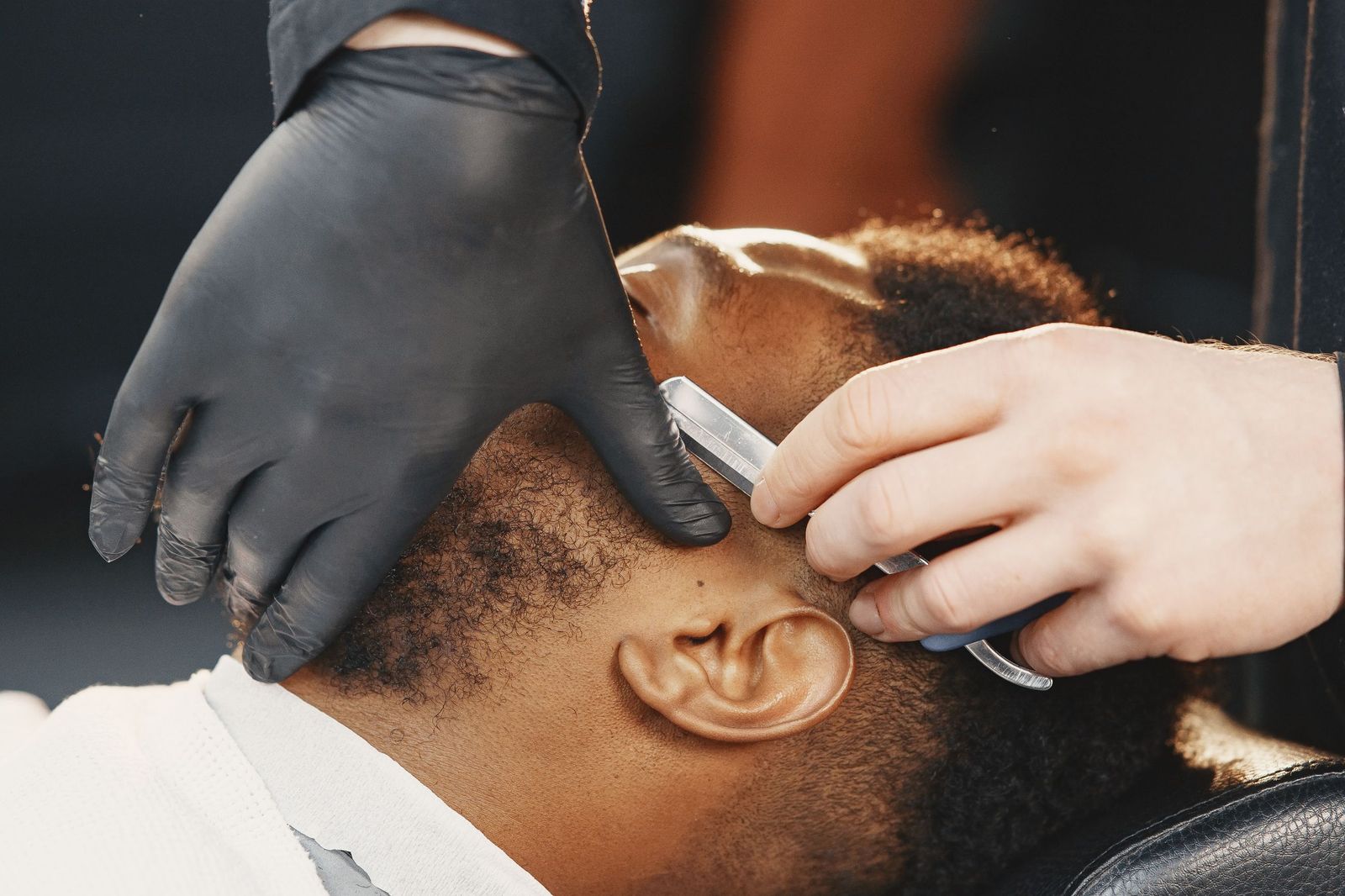 A straight razor at the neckline, the barber’s gloved hand holding the skin flat