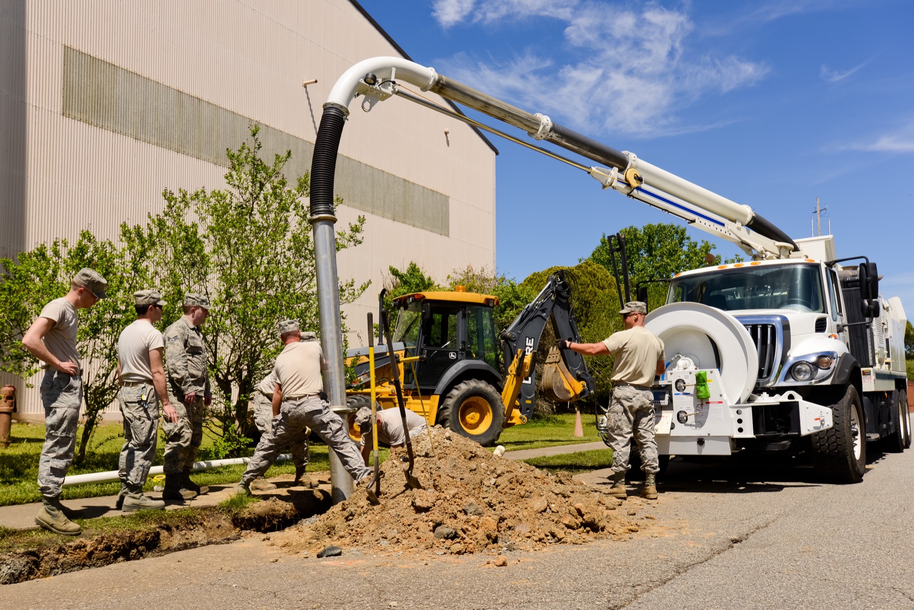 A sewer crew at a dig with a vacuum truck and a backhoe, the boom down into the trench