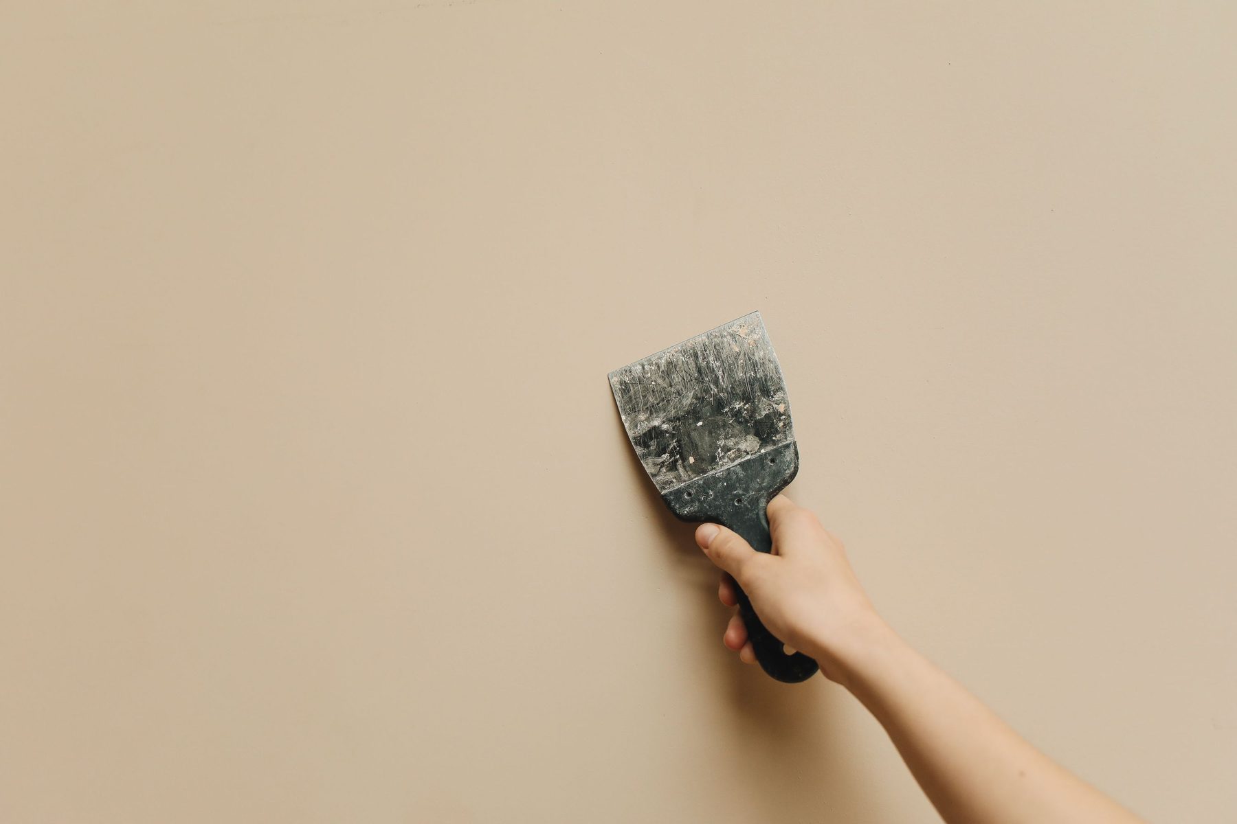 A hand holding a compound-covered putty knife flat against a smooth wall