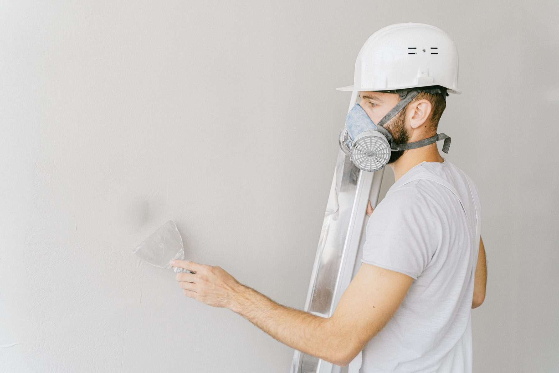 A man in a hard hat and respirator smoothing a patch on a grey wall with a putty knife