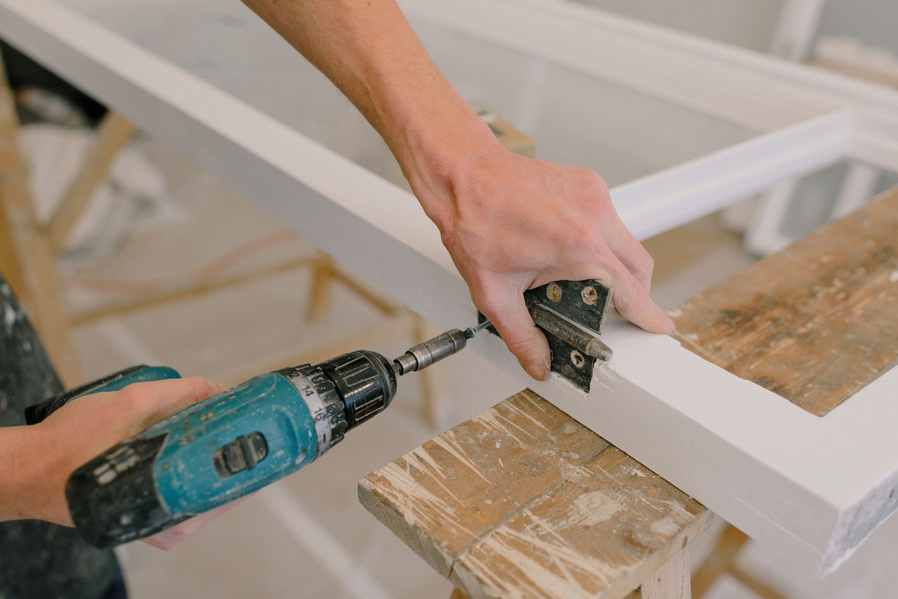 Hands driving a screw through a door hinge into a white-painted frame with a cordless drill