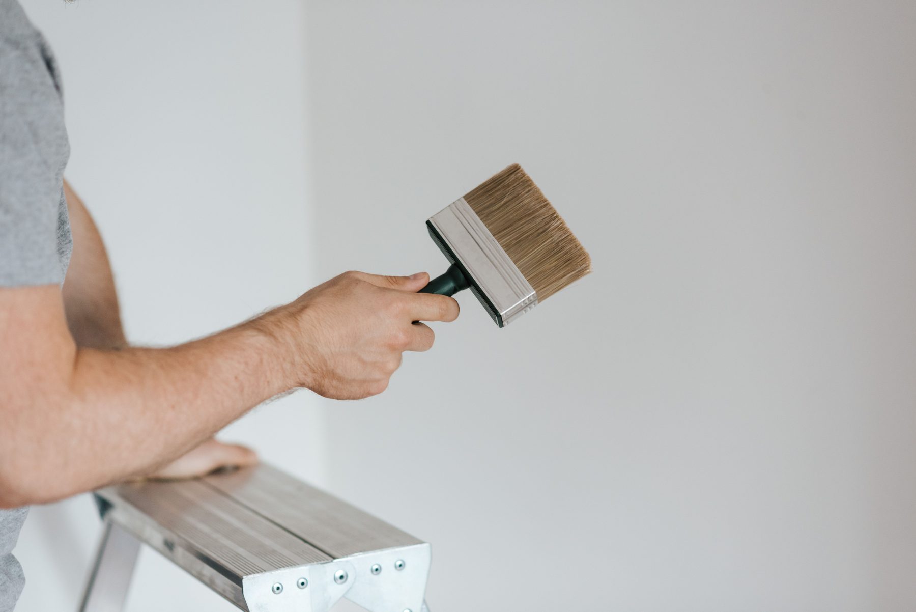 A hand holding a clean wide paintbrush beside a stepladder and a pale wall