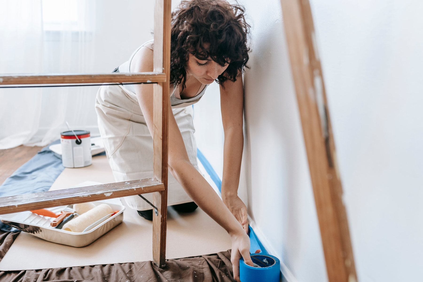 A woman on a drop cloth running painter's tape along a baseboard