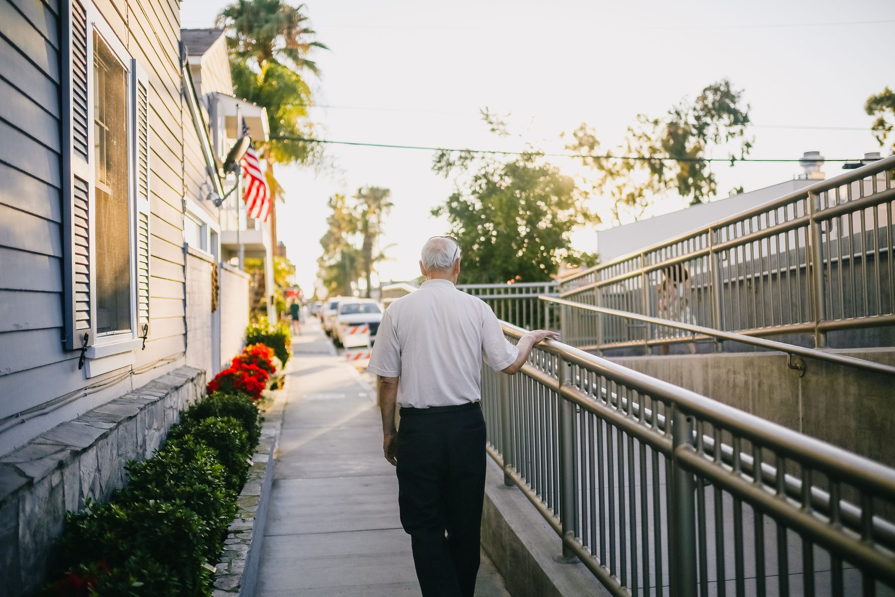 An older man walking a palm-lined sidewalk, hand on a steel rail