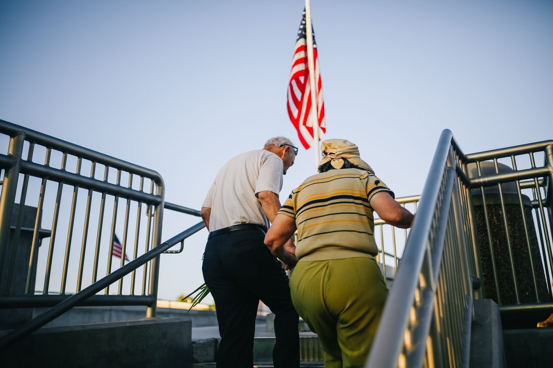 An older couple climbing outdoor steps, one holding the steel handrail