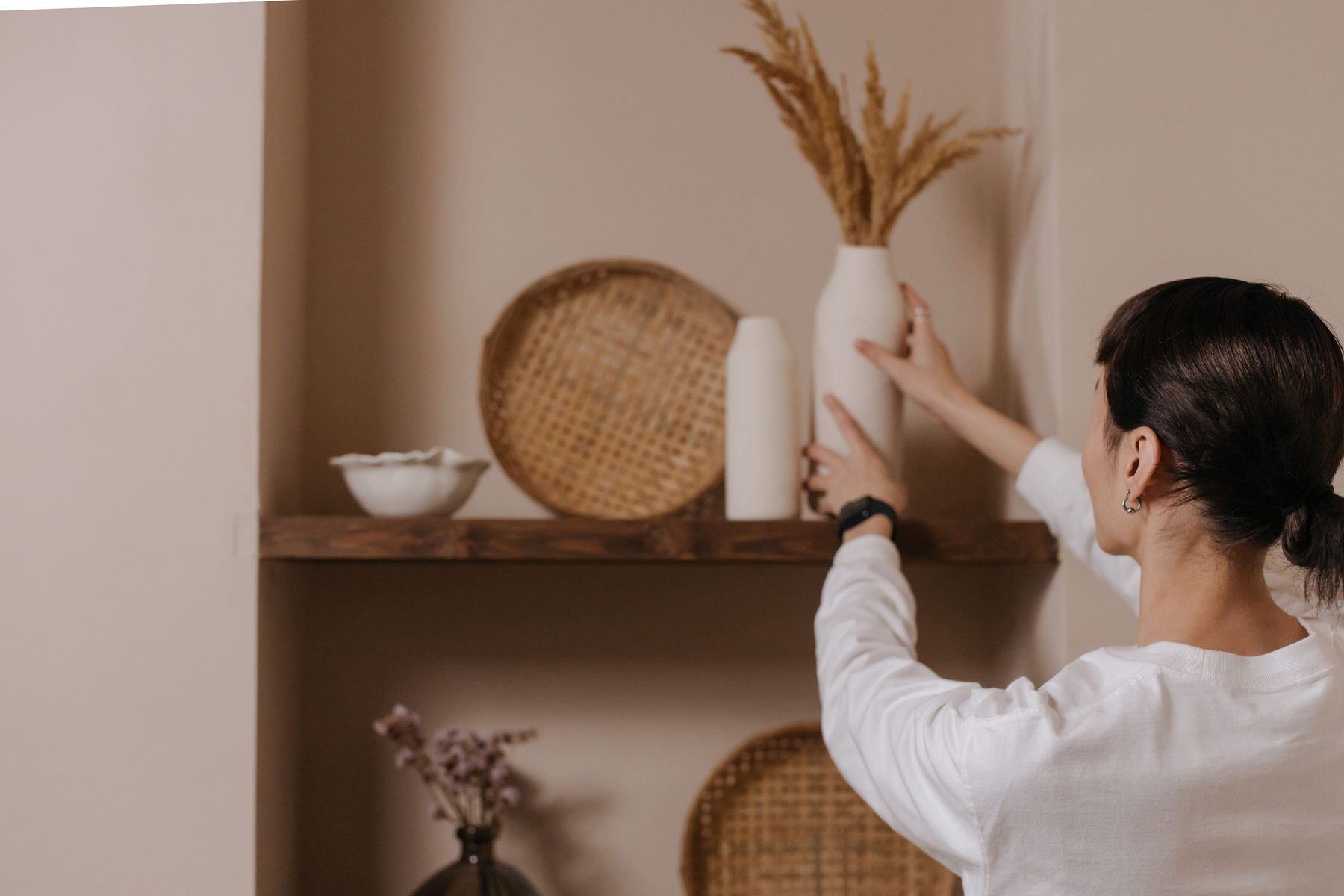 A woman placing a vase of dried grass on a dark floating shelf