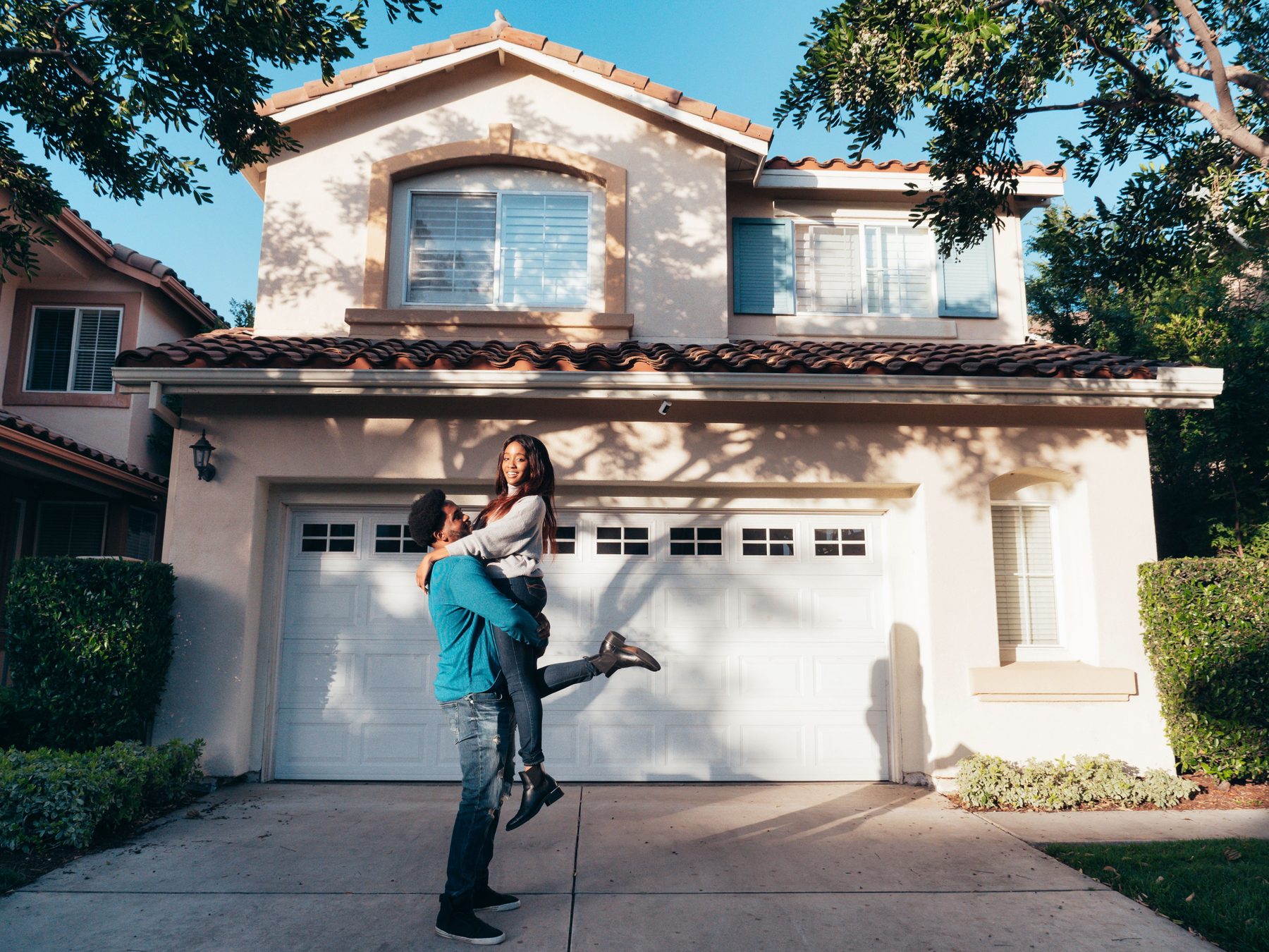 A man lifting a laughing woman in the driveway of a stucco house with a tile roof