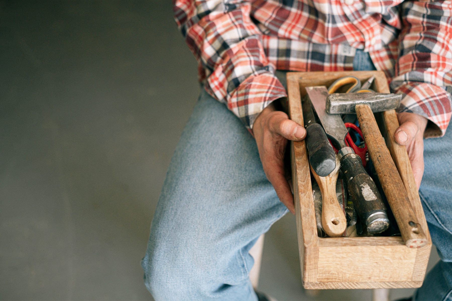 Hands holding an old wooden tool tote with a hammer, chisels and a brush