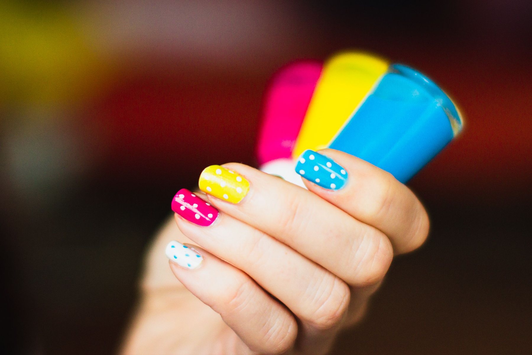Nails painted pink, yellow and blue with white polka dots, the hand holding three polish bottles