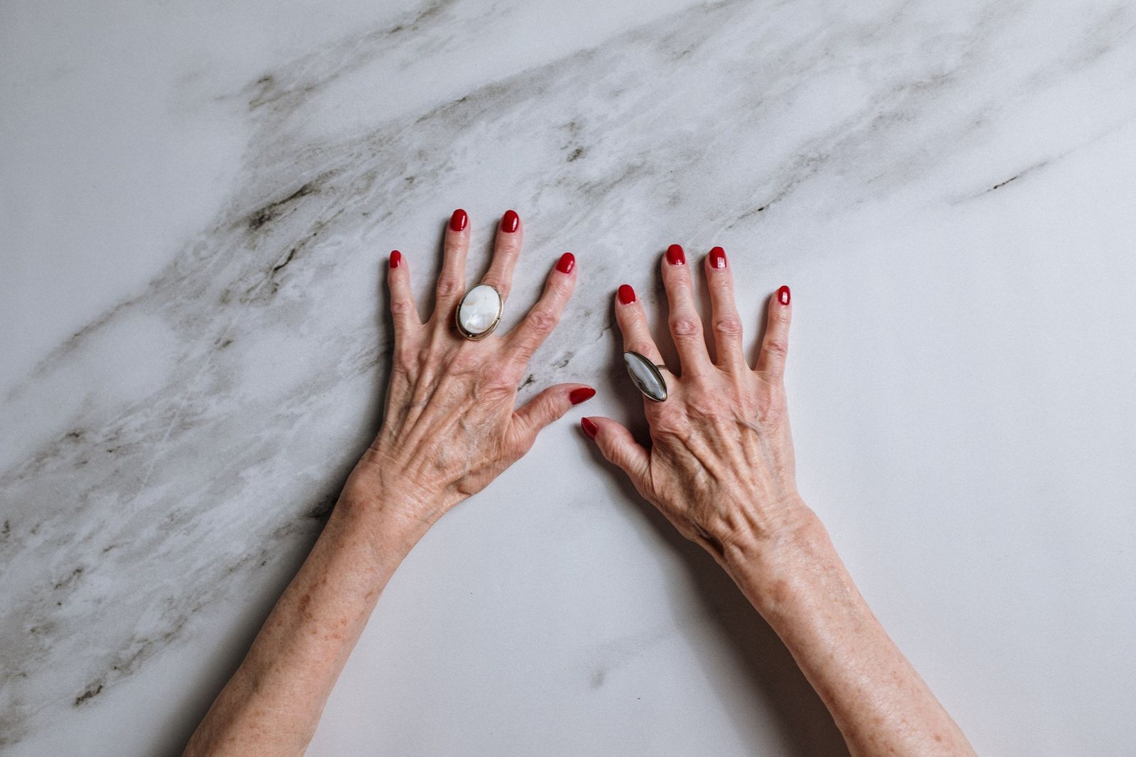 Classic red nails on older hands with statement rings, on white marble