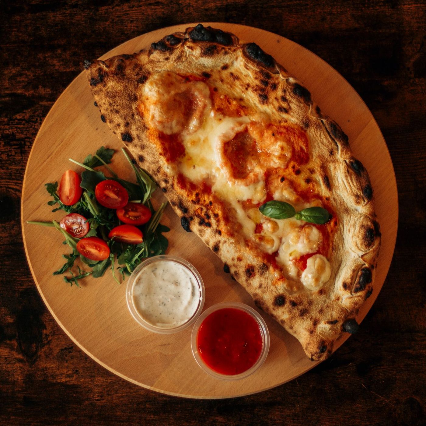 A whole wood-fired calzone with charred crust on a wooden board with salad and marinara dip