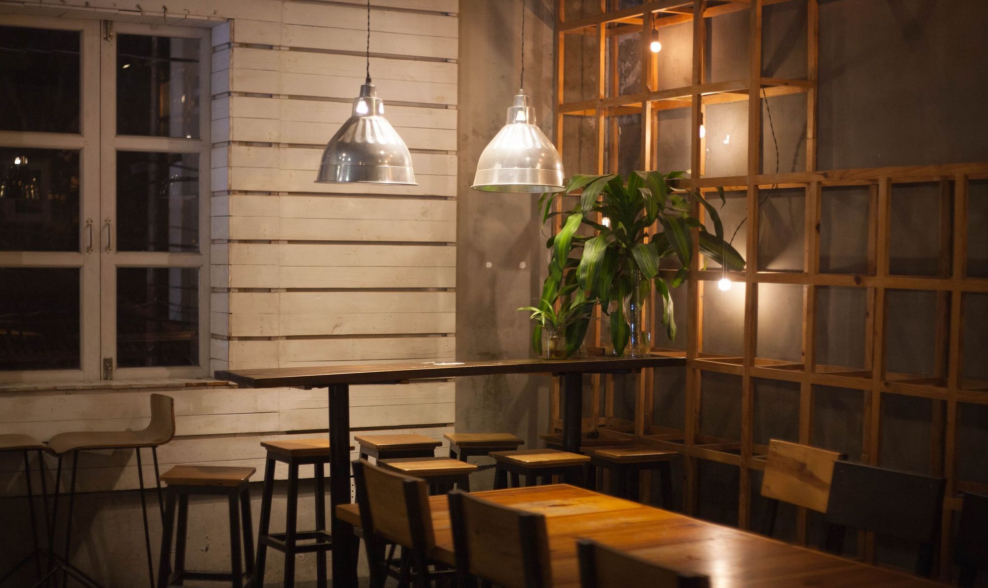 Empty casual restaurant corner at night with wooden tables, bar stools, pendant lamps and a potted plant