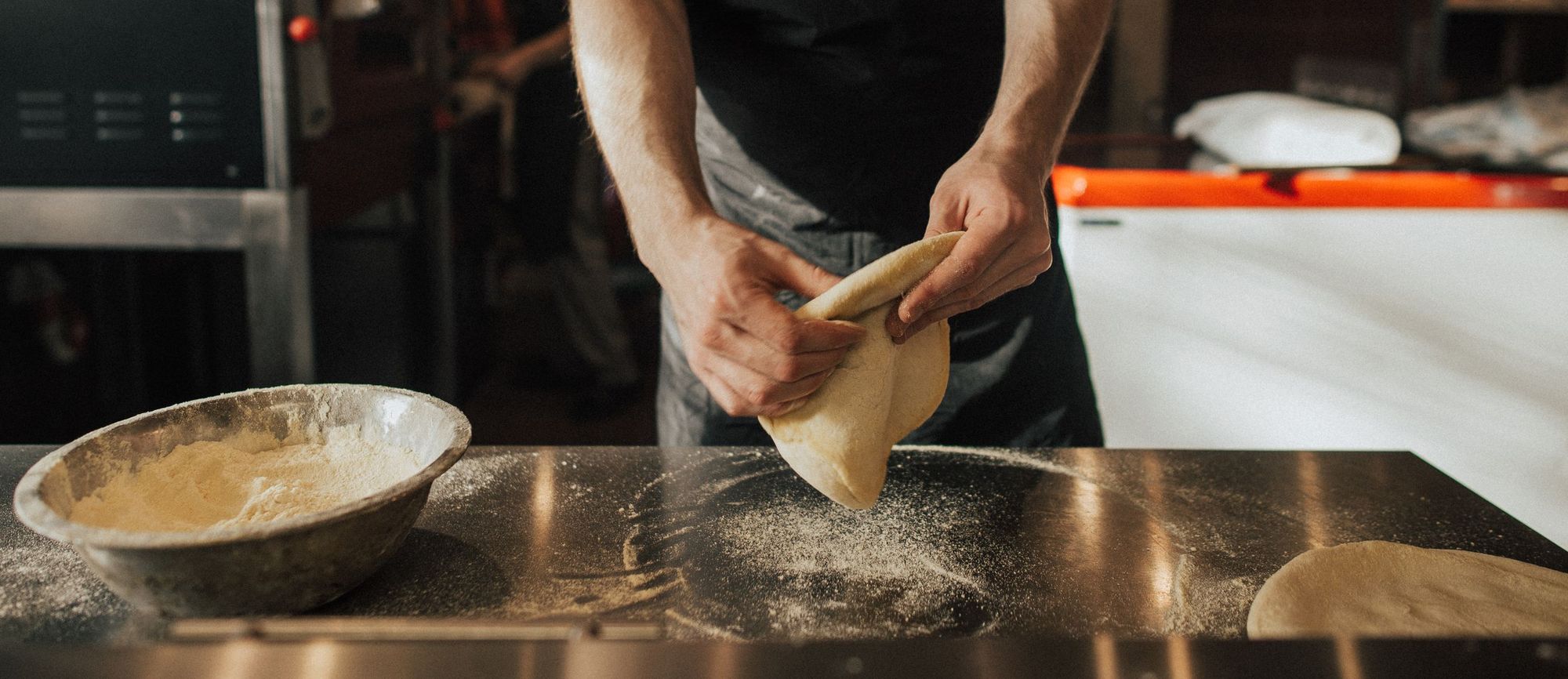 Pizza maker's hands stretching a round of pizza dough over a floured steel counter beside a bowl of flour