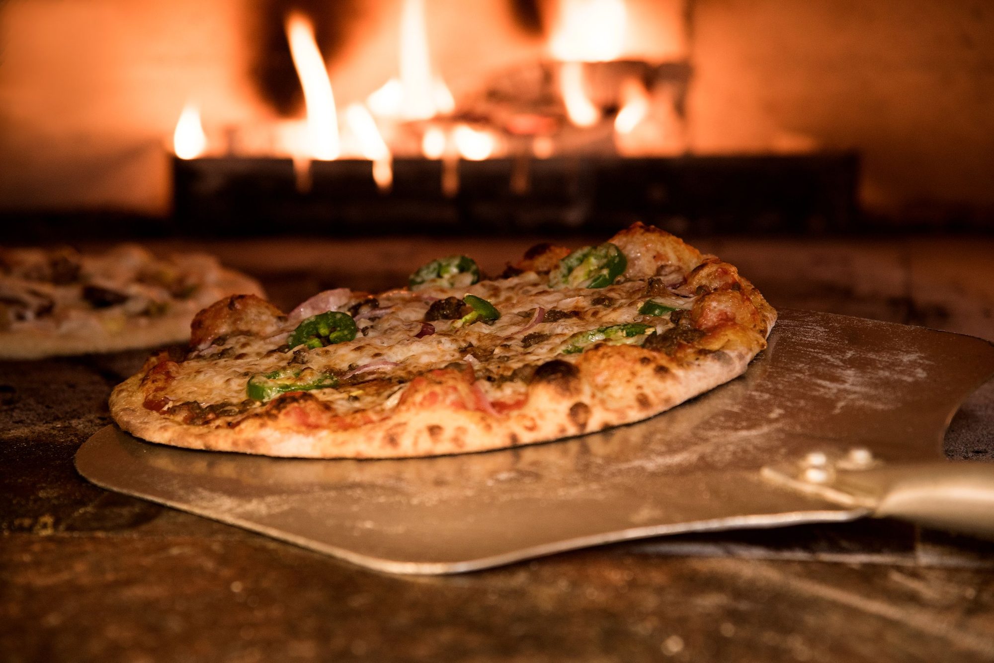 Sausage, jalapeño and red onion pizza on a steel peel at the mouth of a flaming wood-fired oven