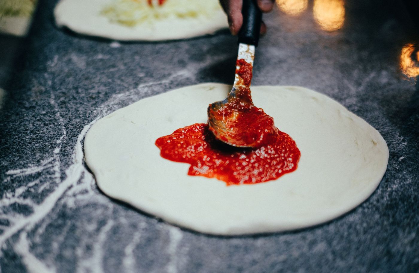 A ladle spreading bright red tomato sauce onto a fresh raw pizza base on a floured stone counter