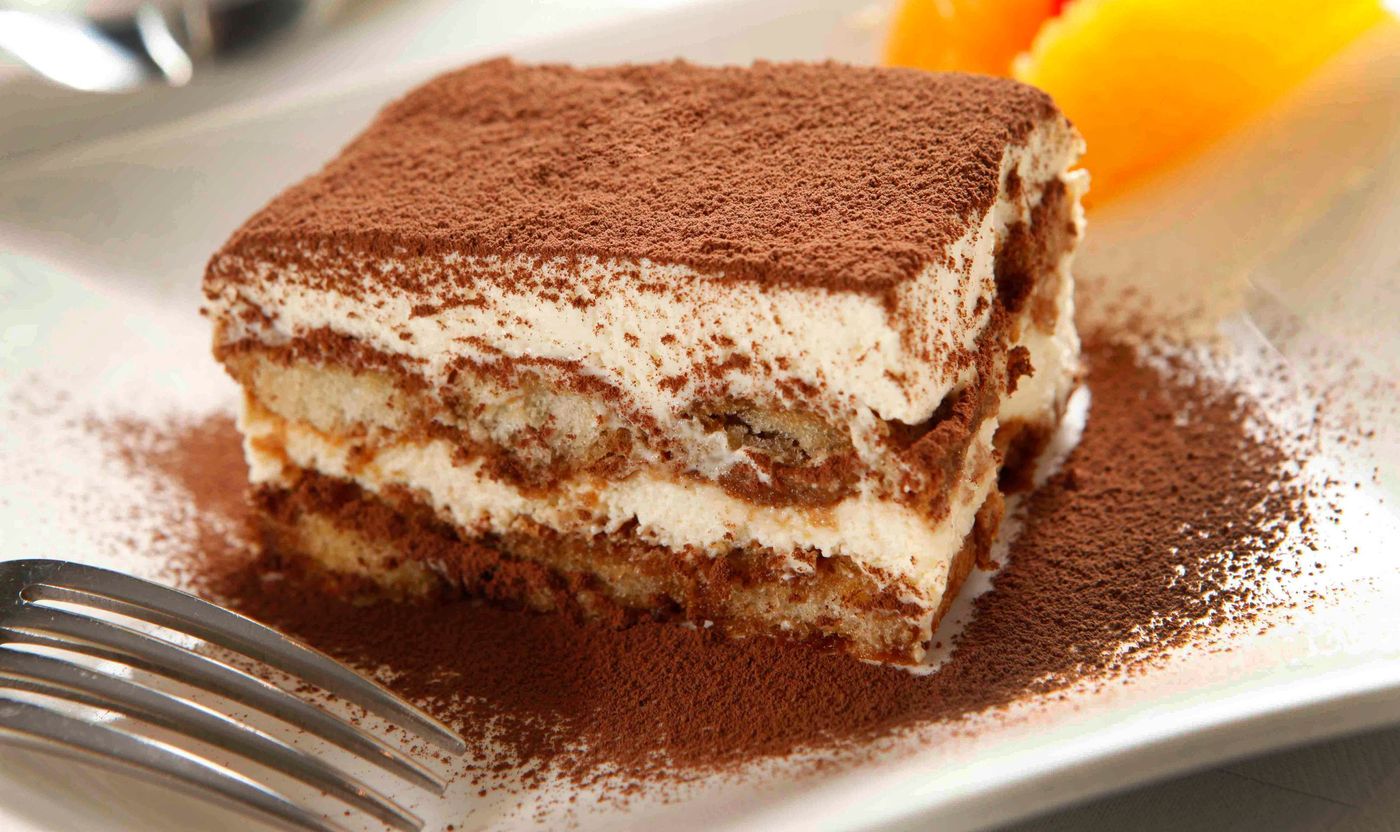 A square slice of tiramisu with ladyfinger layers and a heavy dusting of cocoa on a white plate