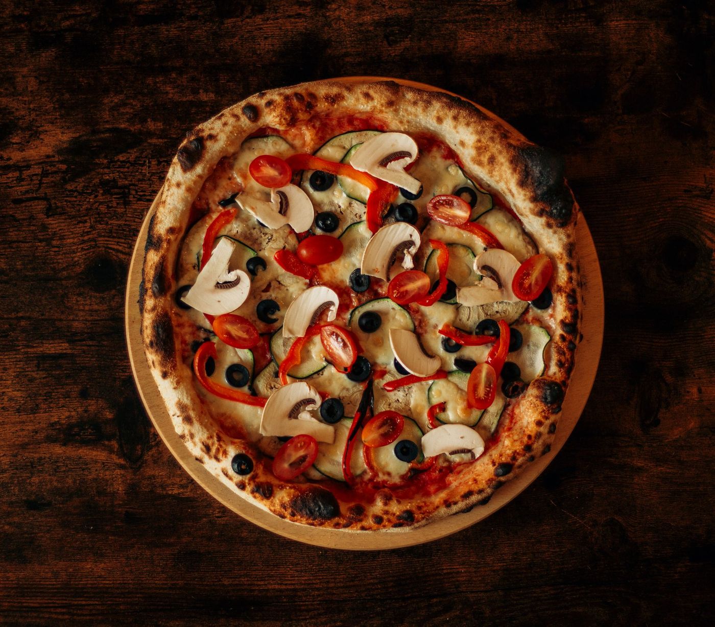 Overhead vegetable pizza with mushrooms, zucchini, red peppers, cherry tomatoes and black olives on dark wood